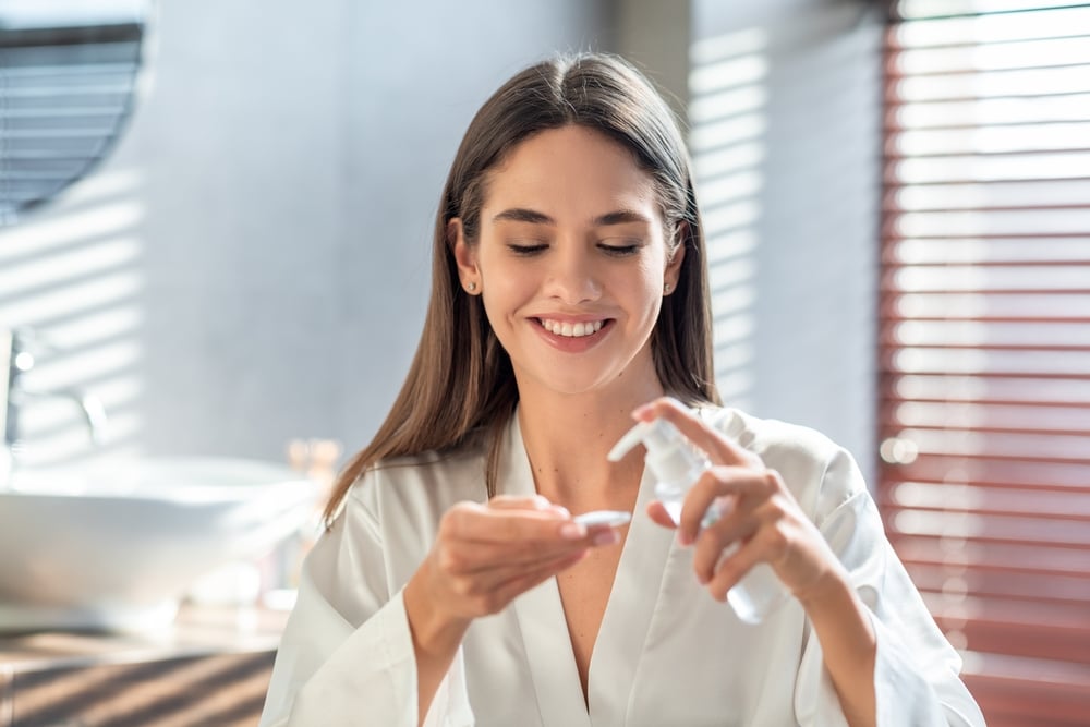 Woman smiling while applying moisturizer to a cotton pad for microneedling aftercare.
