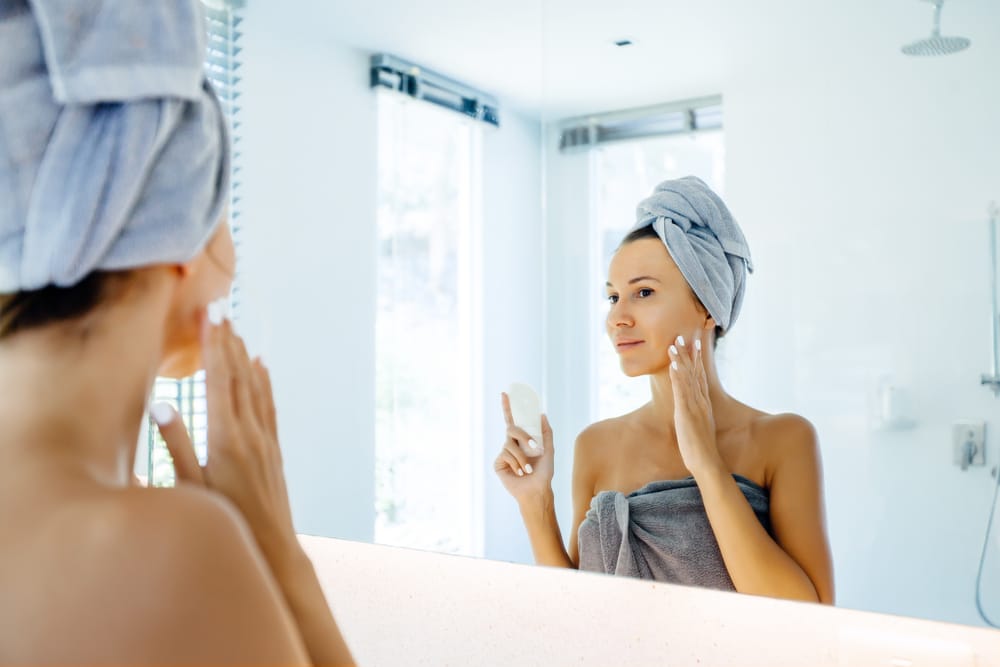 A woman in a towel looking in the mirror and doing skincare for melasma management